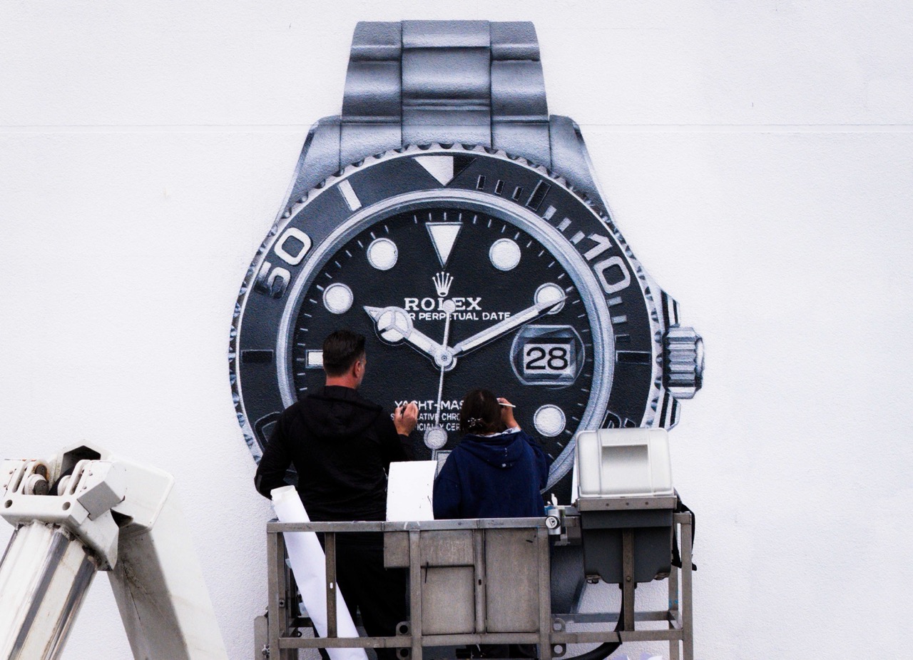 Künstler auf Hebebühne arbeitet präzise an der Form und Schattierung der Rolex Uhr auf einer weißen Wand.