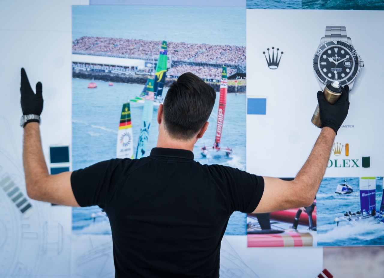 Künstler TANK betrachtet Referenzbilder von Segelrennen und Uhrenmotiven, während er mit Spraydose und Farbproben das Mural vorbereitet.