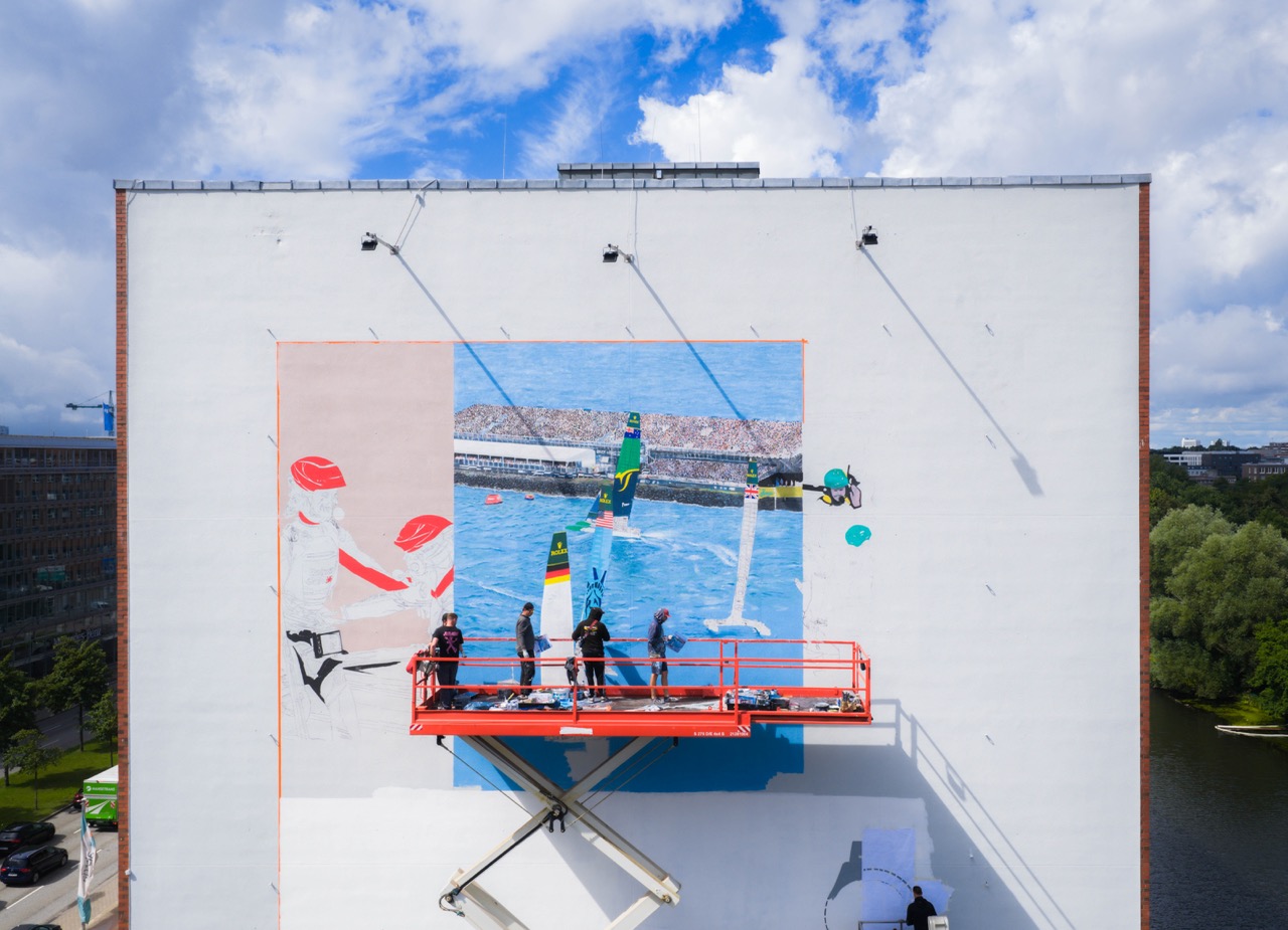 Mehrere Künstler arbeiten auf einer Hebebühne an einem großformatigen Wandbild mit Segelboot-Szene und ersten Skizzen.