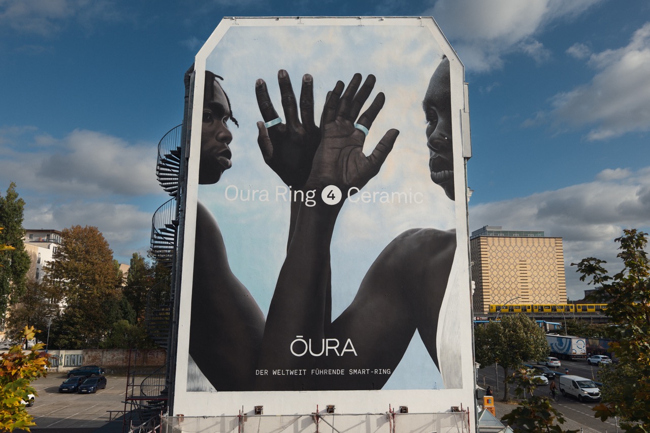 Frontalaufnahme des fertigen Oura Ring 4 Ceramic Murals mit zwei Gesichtern und erhobenen Händen an einer Hauswand in Berlin.
