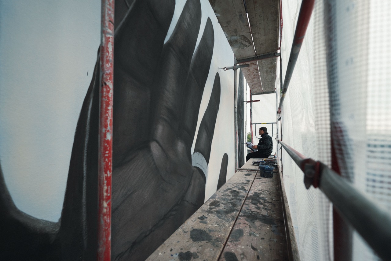 Blick entlang eines Baugerüsts auf ein im Entstehen befindliches Mural mit überdimensionaler Handdarstellung.