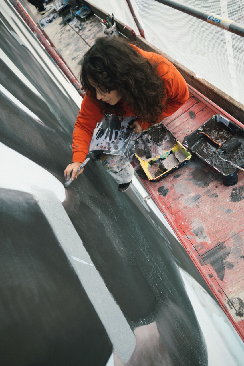 Künstlerin arbeitet auf einem Gerüst an einem großformatigen Mural und trägt mit Pinsel feine Schattierungen auf eine realistische Handdarstellung auf.