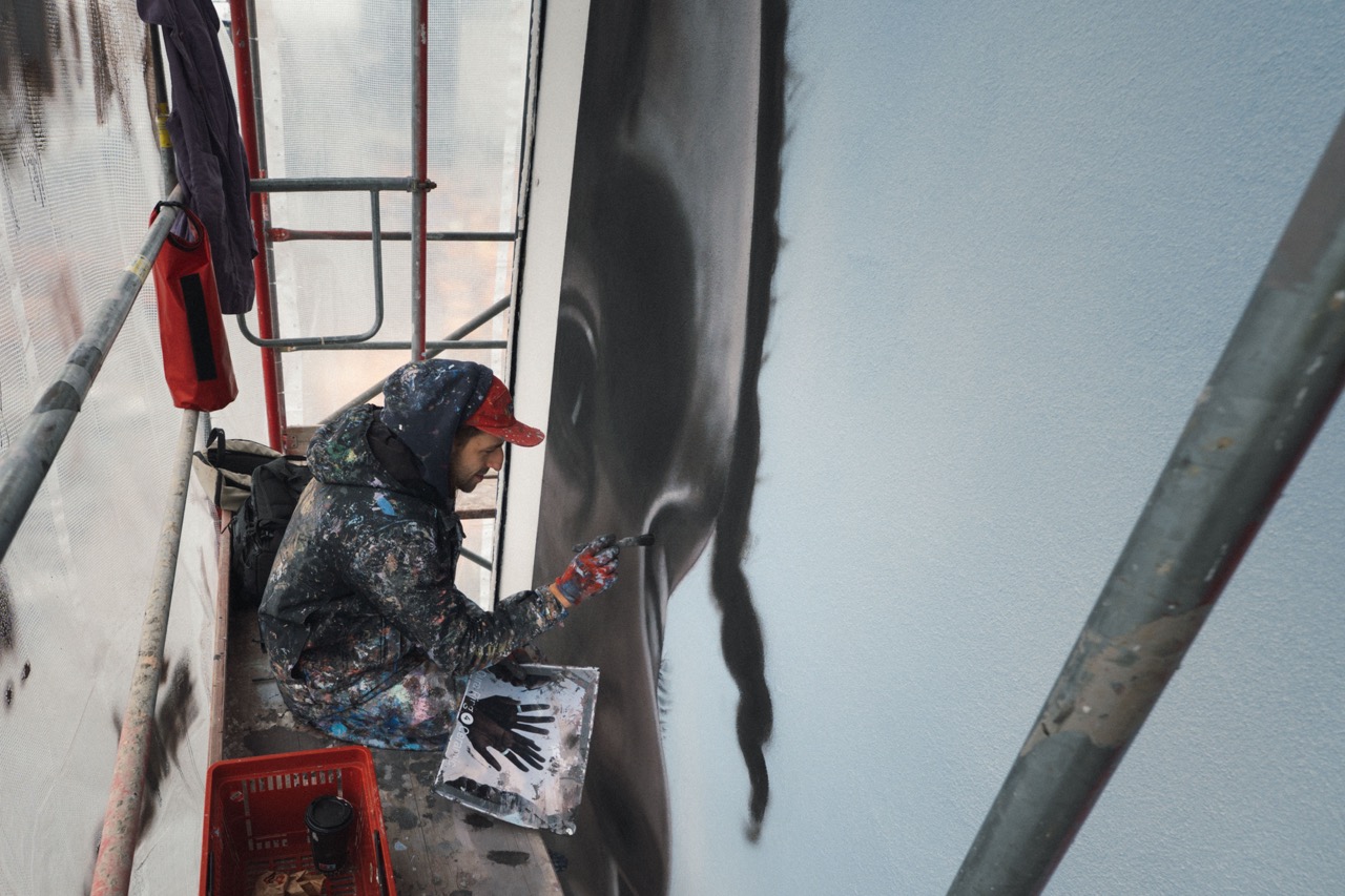 Künstler auf engem Baugerüst malt mit Pinsel die Konturen eines Gesichts für ein großflächiges Wandbild.