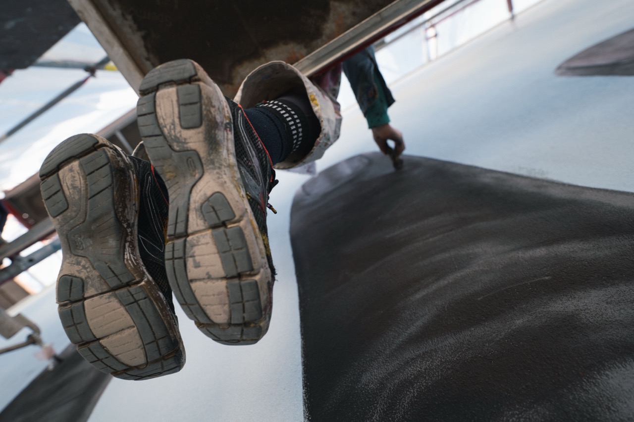 Künstler arbeitet sitzend auf einem Gerüst und malt an einer großflächigen Handdarstellung eines Murals.