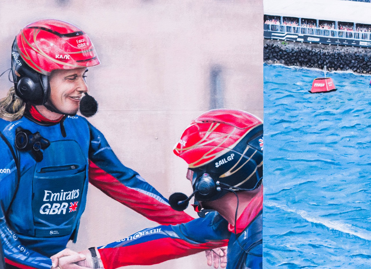 Detail des Murals mit zwei SailGP Athleten in roten Helmen, die sich vor einem Rennen abklatschen.