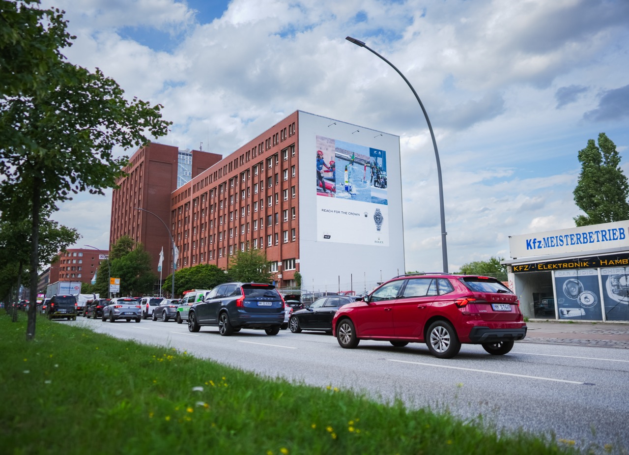Straßenperspektive mit vorbeifahrenden Autos und Blick auf das großflächige Rolex SailGP Mural an einer Gebäudewand.