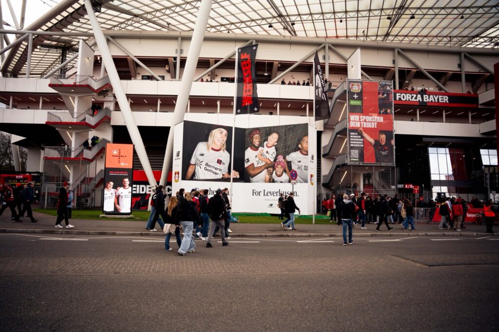 Großformatige Muralinstallation für New Balance und Bayer 04 Leverkusen vor dem Stadion mit mehreren Fassadenelementen, Markeninszenierung und ankommenden Fans am Spieltag.