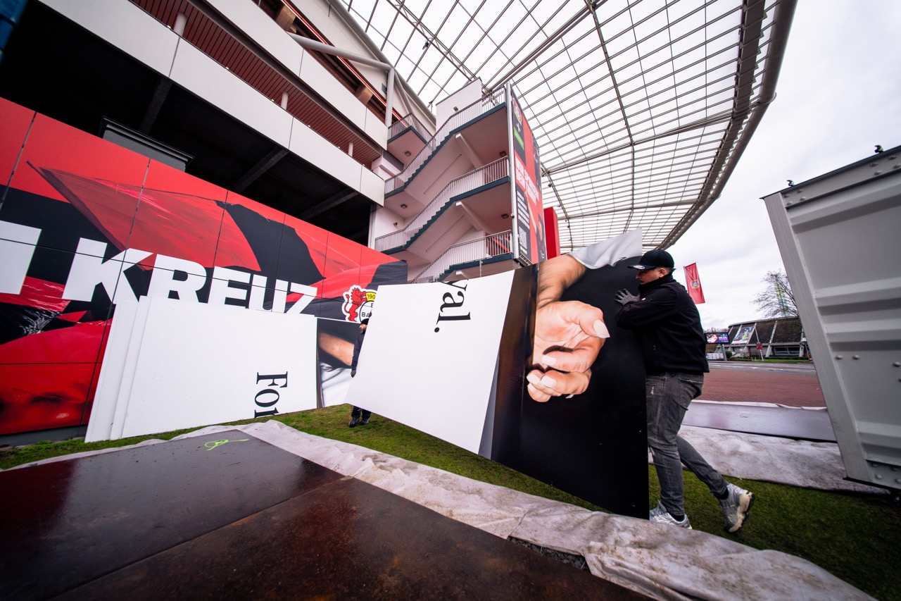 Aufbau der großformatigen Muralinstallation vor dem Stadion von Bayer 04 Leverkusen mit montierten Bildmodulen und Team bei der Ausrichtung.