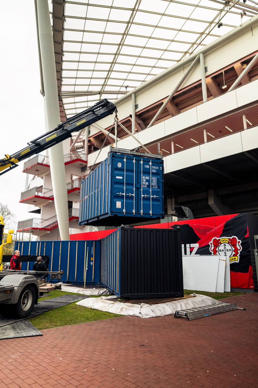 Containerinstallation während des Aufbaus vor dem Stadion mit Kran und vorbereiteten Modulen für die Markenaktivierung.