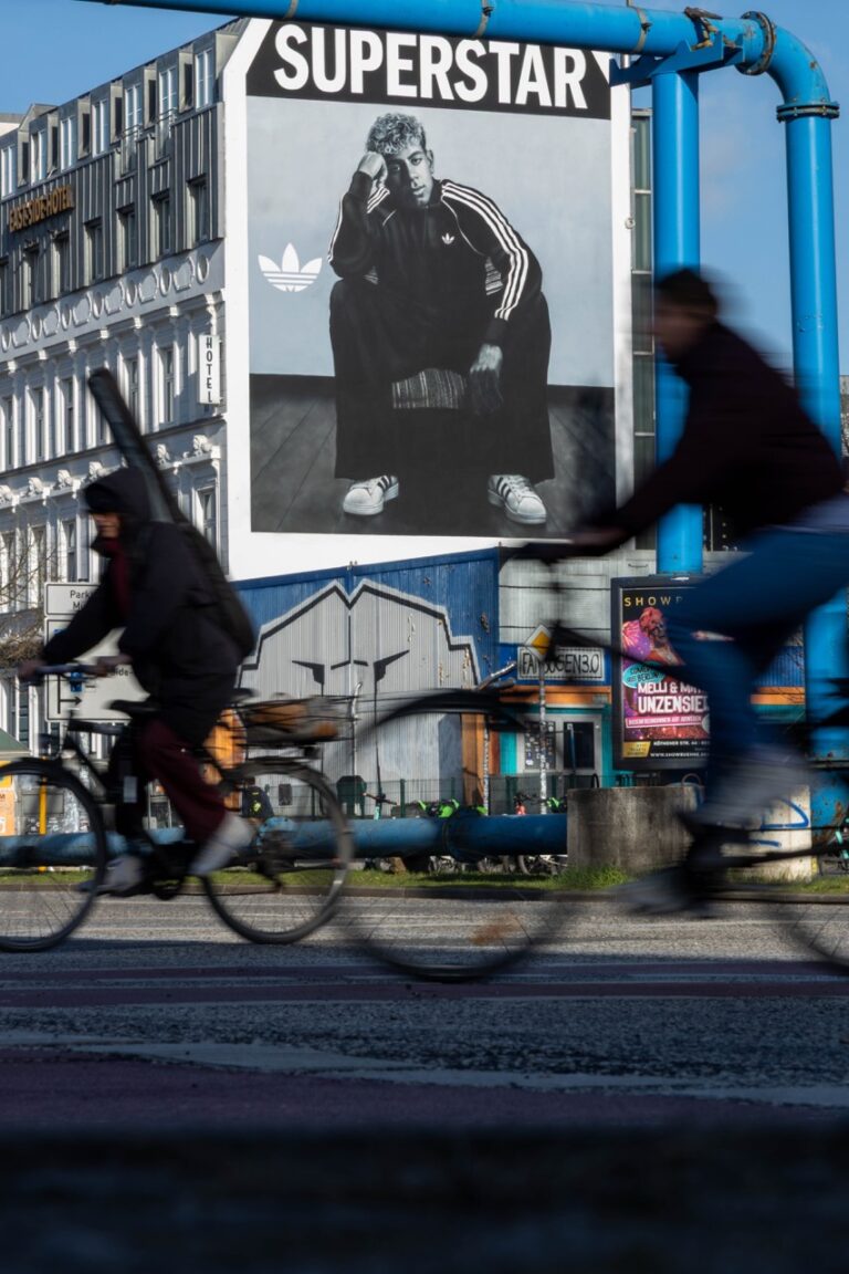 Großformatiges adidas Originals Mural mit Fußballstar Lamine Yamal an der Berliner East Side Gallery. Im Vordergrund fahren verschwommen Radfahrer vorbei, während das Schwarz-Weiß-Motiv für den adidas Superstar über dem Berliner Straßenverkehr sichtbar ist.