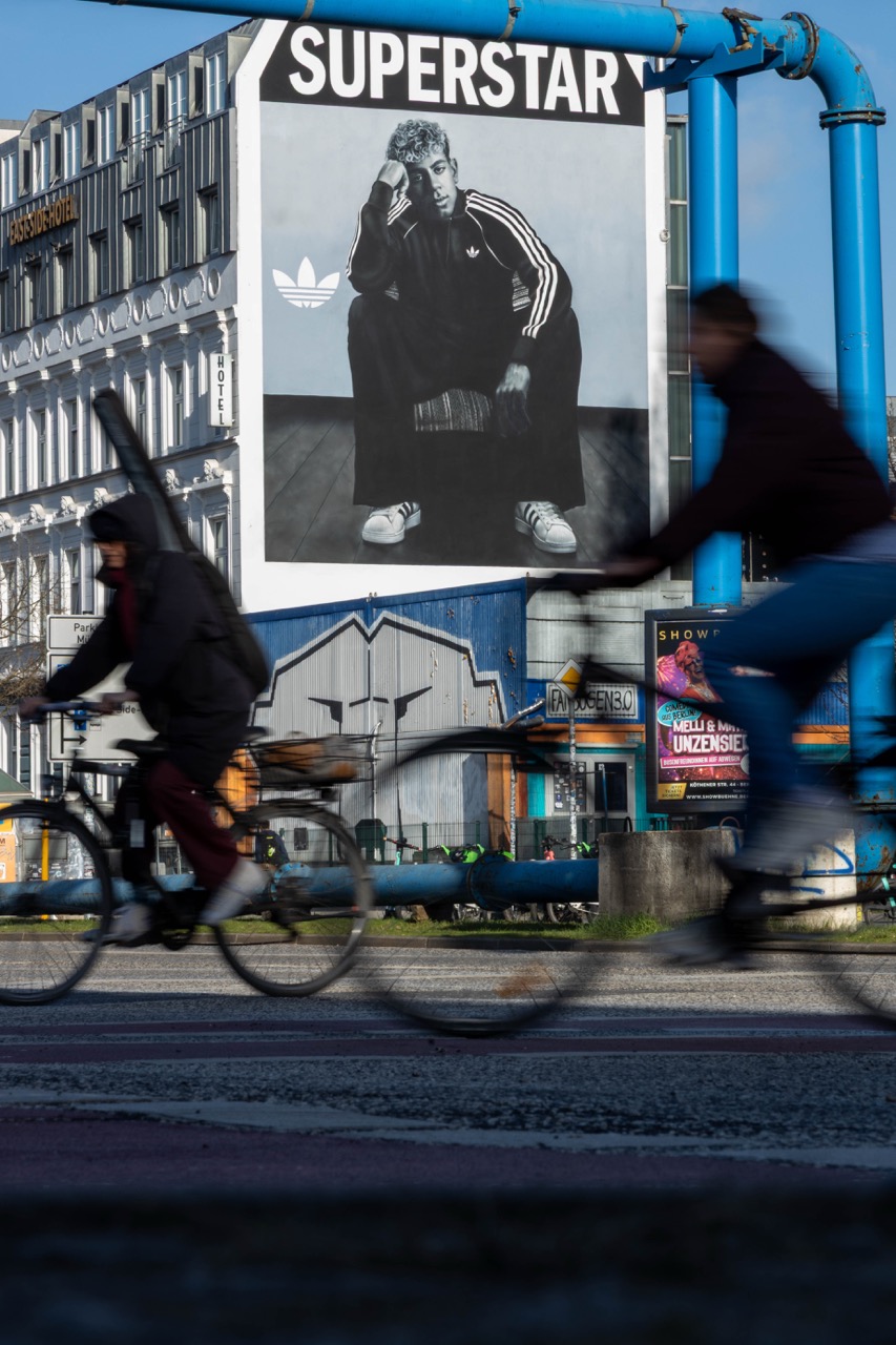 Großformatiges adidas Originals Mural mit Fußballstar Lamine Yamal an der Berliner East Side Gallery. Im Vordergrund fahren verschwommen Radfahrer vorbei, während das Schwarz-Weiß-Motiv für den adidas Superstar über dem Berliner Straßenverkehr sichtbar ist.