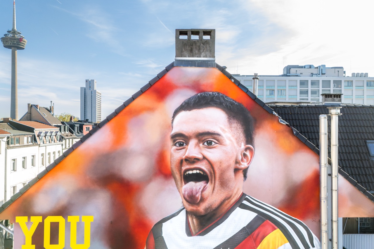 Nahaufnahme des fertigen Florian-Wirtz-Murals vor orangefarbenem Hintergrund mit Kölner Fernsehturm im Hintergrund.
