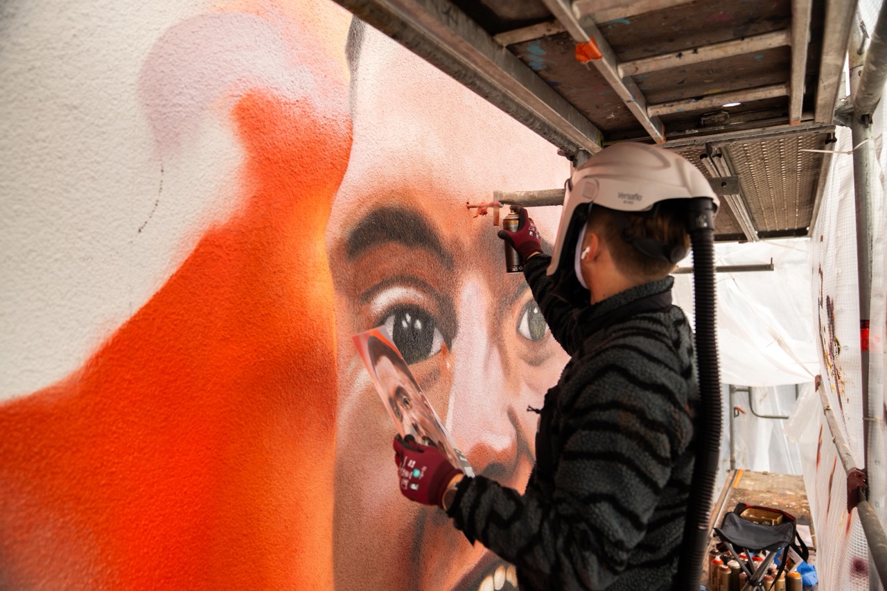 Artist sprüht Details am Gesicht von Florian Wirtz auf die Hauswand und hält dabei die Referenzvorlage des DFB Trikots in der Hand.