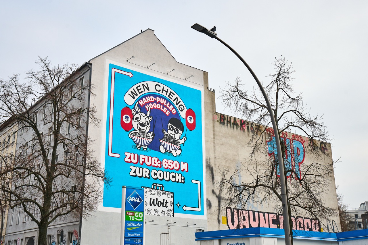 Großformatiges WOLT-Mural an einer Hausfassade in Berlin-Kreuzberg mit Comicfiguren, Wen Cheng Noodles Branding und Wegweisung zur Bestellung per Lieferdienst.