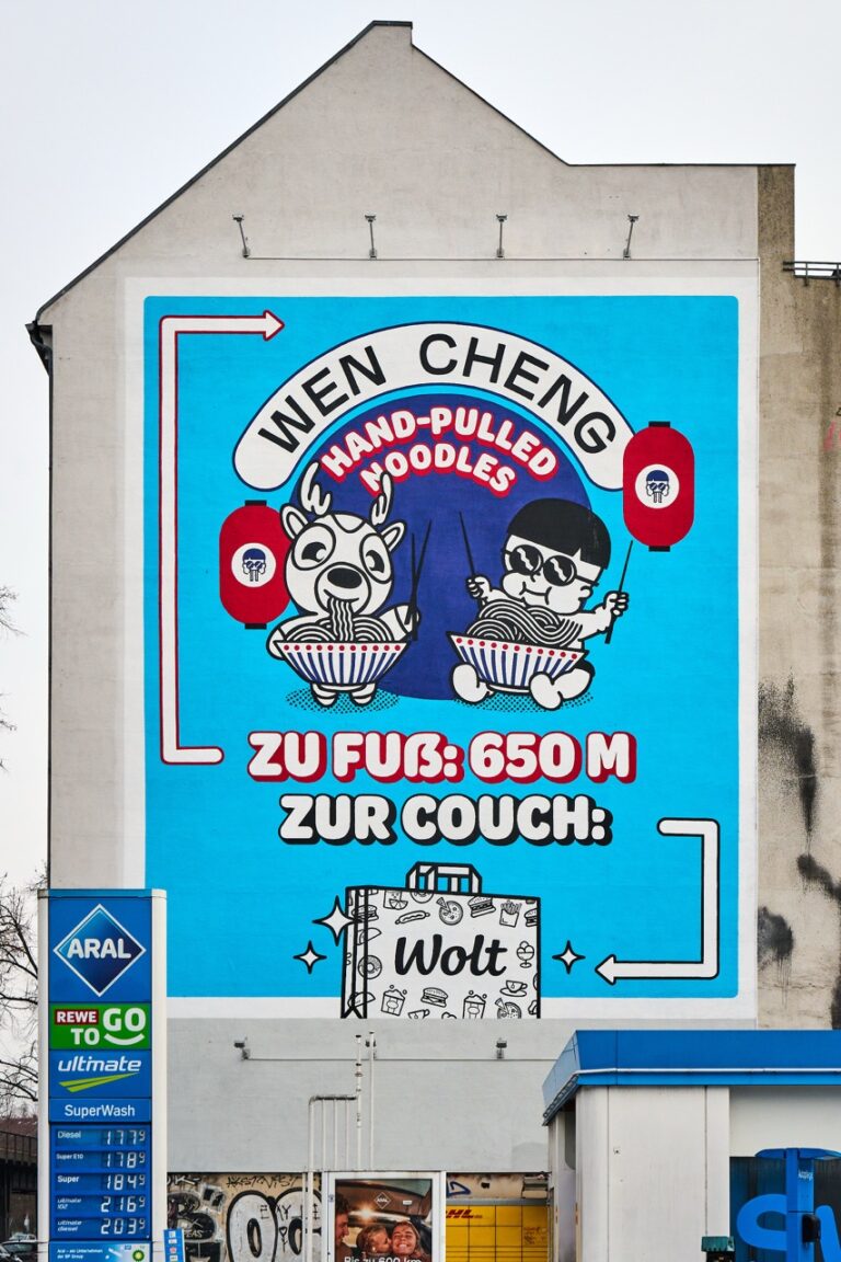 Großformatiges WOLT-Mural an einer Hausfassade in der Skalitzer Straße in Berlin-Kreuzberg. Das Artwork zeigt zwei Comicfiguren beim Nudelessen vor blauem Hintergrund mit dem Schriftzug „WEN CHENG – Hand-Pulled Noodles“. Pfeile weisen auf die Entfernung zum Restaurant sowie zur Bestellung über WOLT hin.