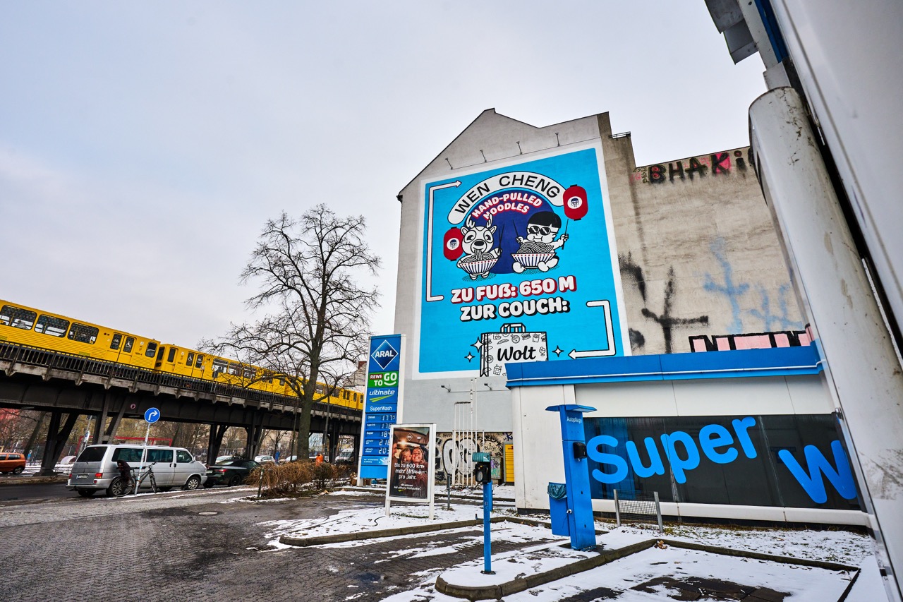 WOLT-Mural in Berlin-Kreuzberg neben einer Hochbahnstrecke der U-Bahn und einer ARAL-Tankstelle im winterlichen Stadtbild.