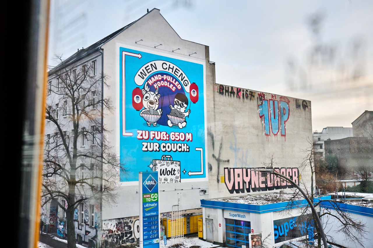 Blick aus einem Fenster auf das großformatige WOLT-Mural an einer Hauswand in Berlin-Kreuzberg mit angrenzenden Graffiti-Fassaden.