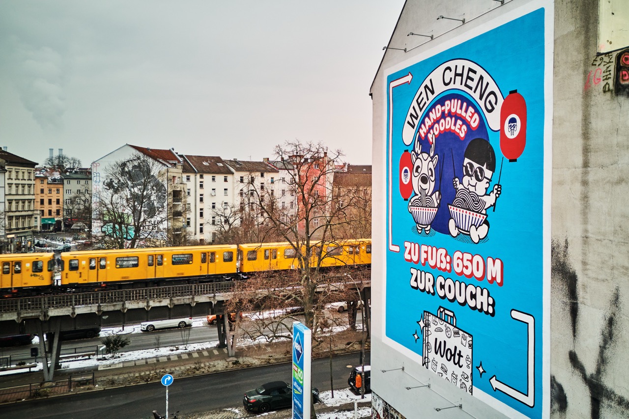 Seitliche Ansicht des WOLT-Murals mit vorbeifahrender gelber Berliner U-Bahn im Hintergrund.