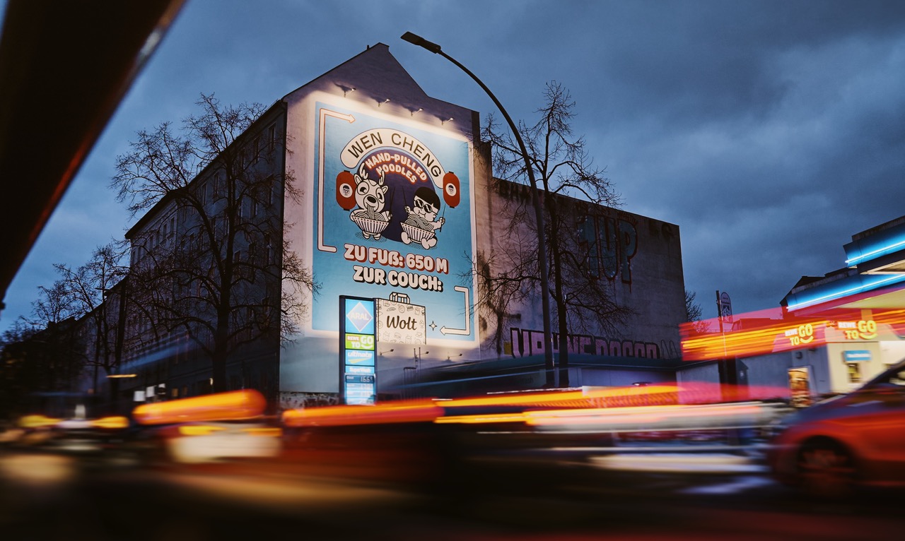 Beleuchtetes WOLT-Mural bei Nacht in Berlin-Kreuzberg mit Lichtspuren vorbeifahrender Autos im Vordergrund.