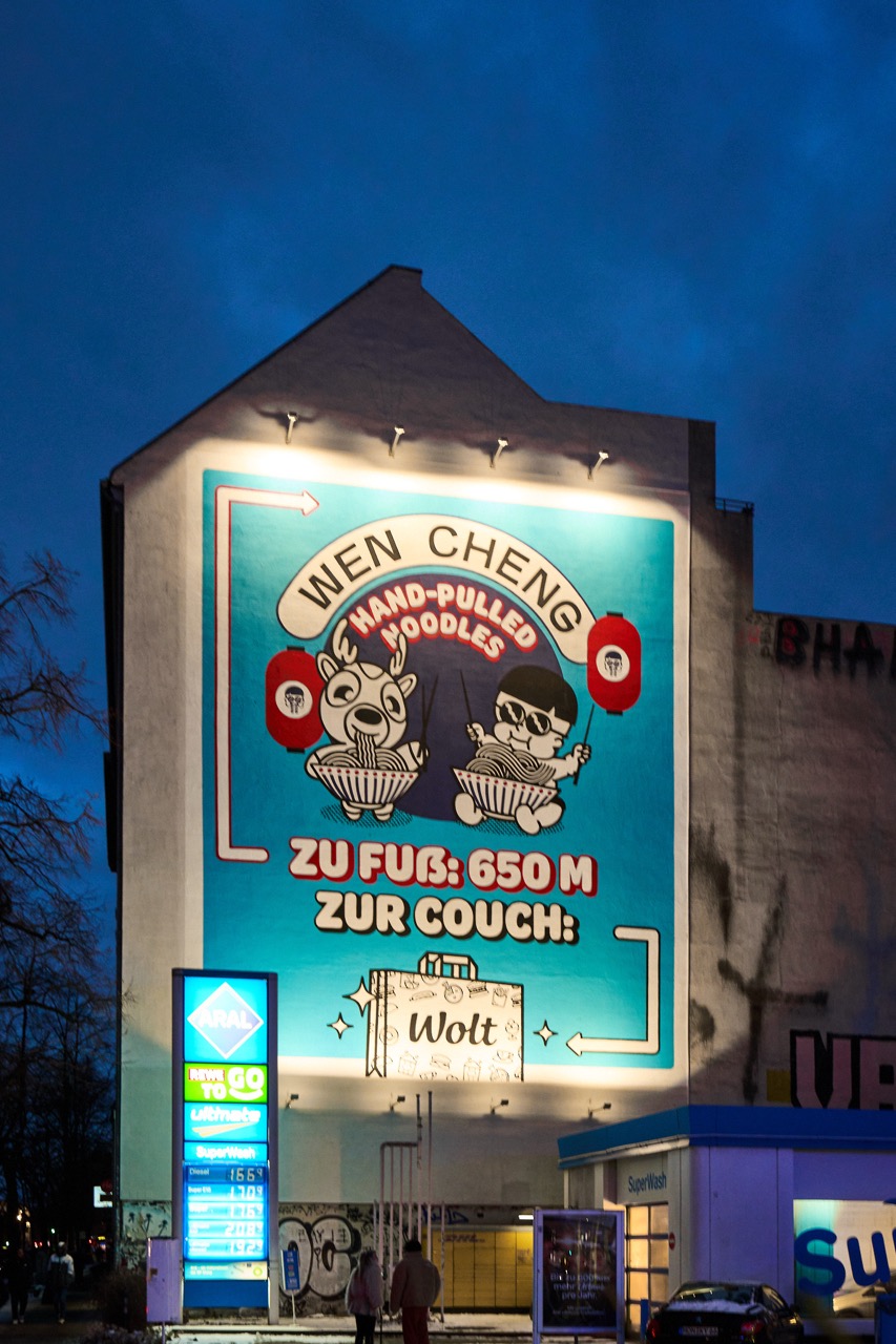 Nachts beleuchtetes WOLT-Mural an einer Hausfassade in Berlin-Kreuzberg mit leuchtender ARAL-Stele im Vordergrund.