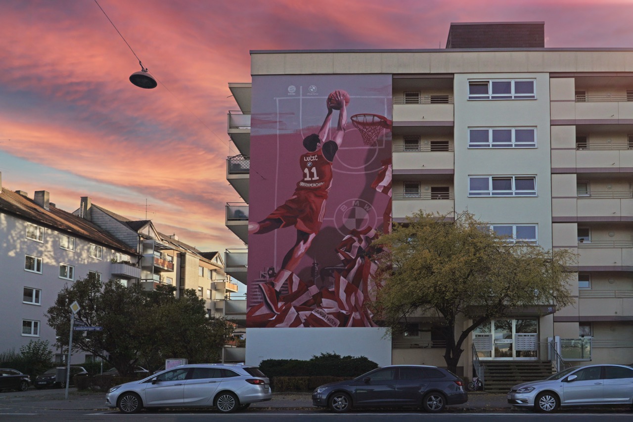 Frontale Aufnahme des FC Bayern Basketball Murals an einer Wohnhausfassade mit pinkfarbenem Himmel im Hintergrund.