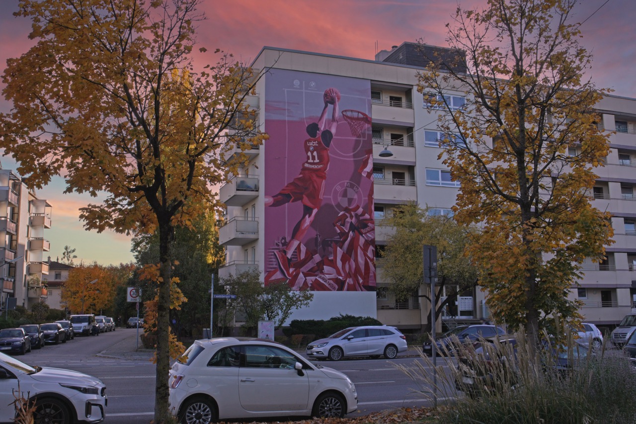 Großformatiges FC Bayern Basketball Mural mit Spieler in Dunking-Position an einer Münchner Hausfassade bei Sonnenuntergang.