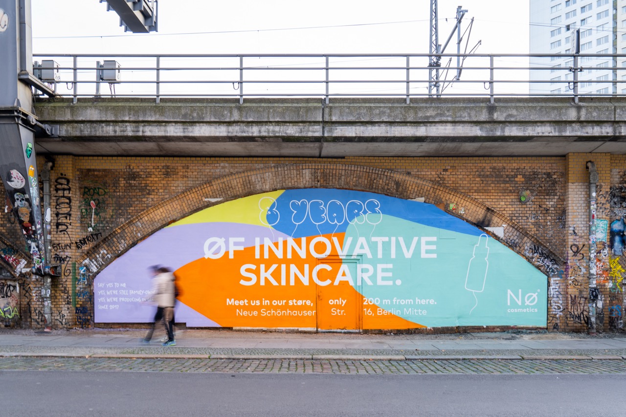 Großflächige Nø Cosmetics Kampagne unter einer Berliner Bahnbrücke in der Dircksenstraße nahe Alexanderplatz mit buntem Design, typografischer Botschaft und vorbeilaufender Person.