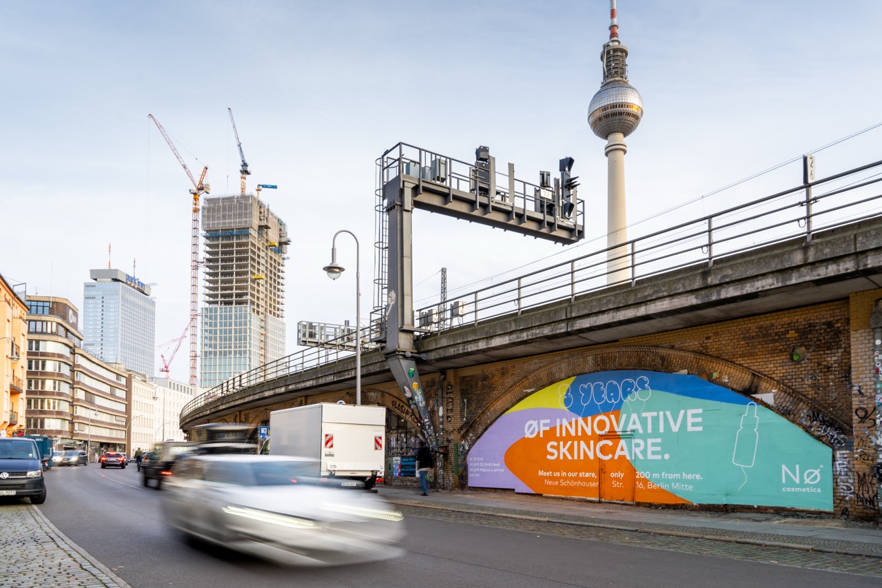 Weitwinkelaufnahme der Nø Cosmetics Installation an der Dircksenstraße in Berlin mit Fernsehturm, Bahntrasse und vorbeifahrenden Autos im Abendlicht.