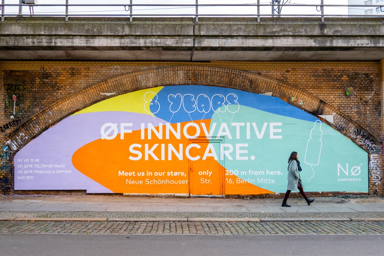 Frontale Aufnahme der großformatigen Nø Cosmetics Kampagne unter der Bahnbrücke in Berlin mit vorbeilaufender Passantin und urbanem Streetart-Umfeld.