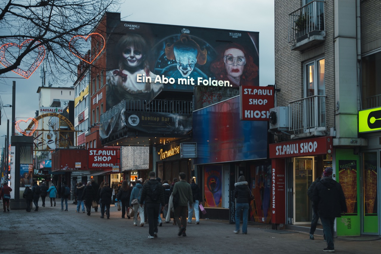 HBO-Max-Mural „Ein Abo mit Folgen“ über den Straßen von St. Pauli in Hamburg mit beleuchtetem 3D-Schriftzug und Passanten im Abendlicht.