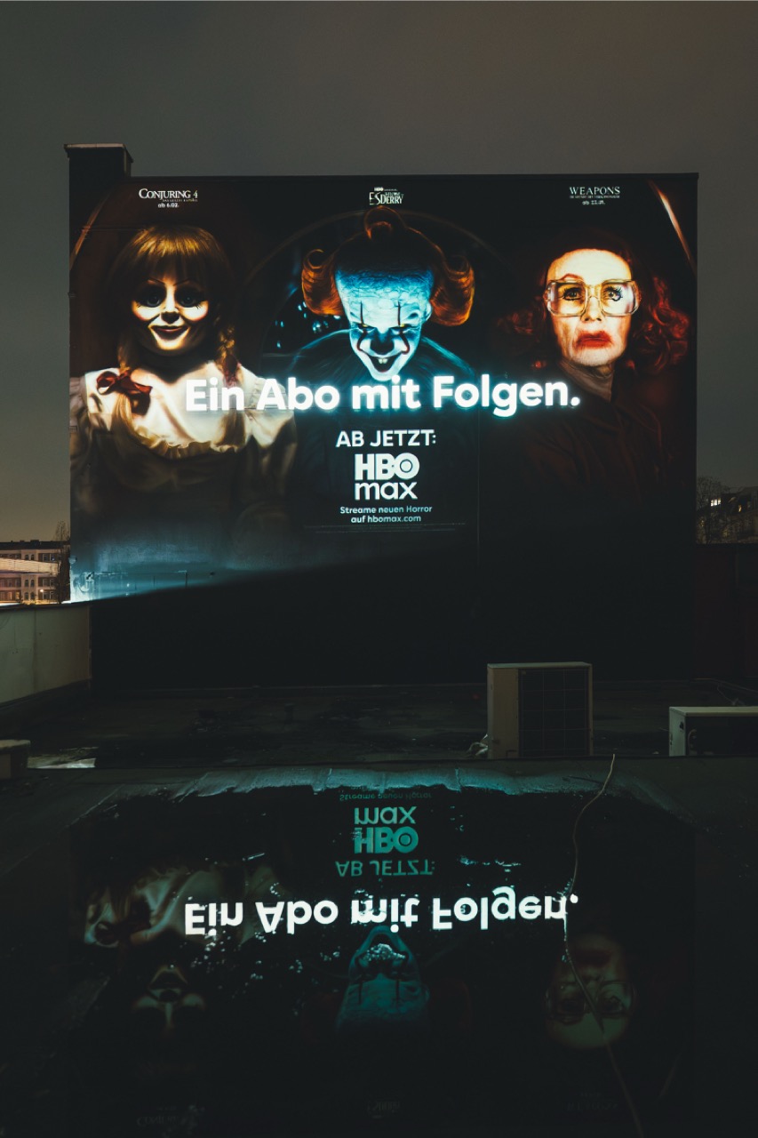 HBO-Max-Mural bei Nacht mit Spiegelung des beleuchteten Schriftzugs auf einer regennassen Dachfläche.