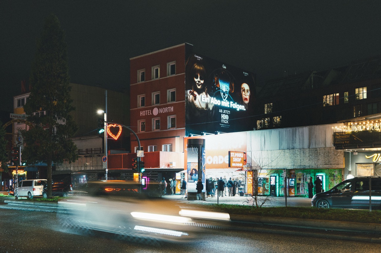 HBO-Max-Mural an einer Gebäudefassade in Hamburg bei Nacht mit Lichtspuren vorbeifahrender Fahrzeuge im Vordergrund.