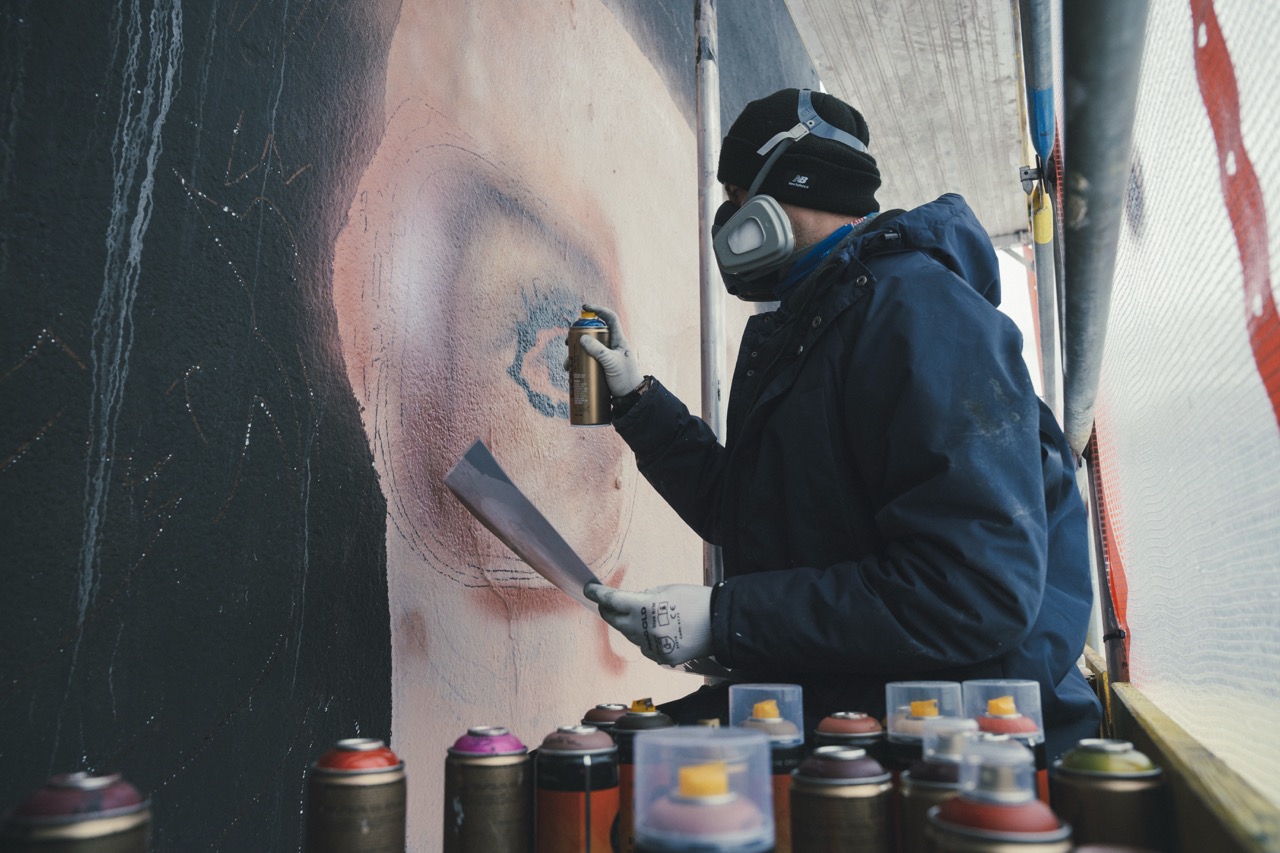 Künstler arbeitet mit Spraydose und Referenzvorlage an einem großformatigen Portrait des HBO-Max-Murals.
