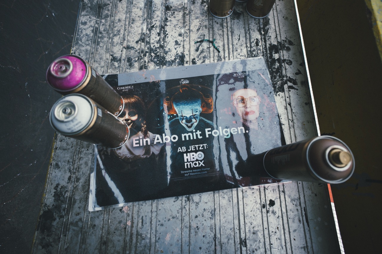 Ausgedruckte Motivvorlage der HBO-Max-Kampagne „Ein Abo mit Folgen“ zwischen Sprühdosen auf einer mit Farbresten bedeckten Arbeitsplattform.
