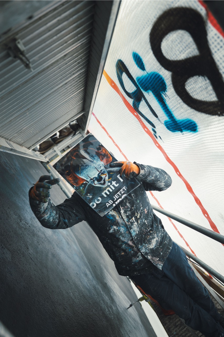 Künstler hält eine Motivvorlage mit Horrorfigur vor das Gesicht und posiert auf dem Gerüst des HBO-Max-Murals.
