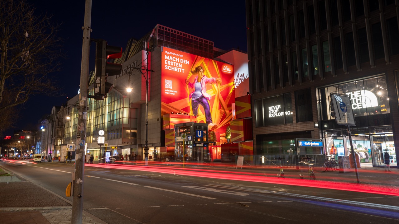 Weitwinkelaufnahme des beleuchteten Lilly Murals mit Lichtspuren des Verkehrs am Kurfürstendamm.