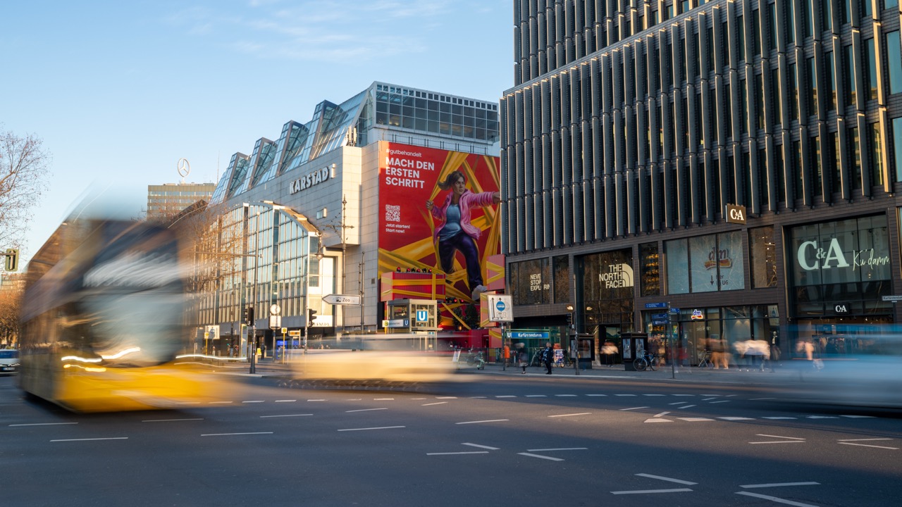 Großflächige Außenansicht des Lilly Murals am Ku’damm mit vorbeiziehendem Verkehr und Blick auf die gesamte Fassadeninszenierung.