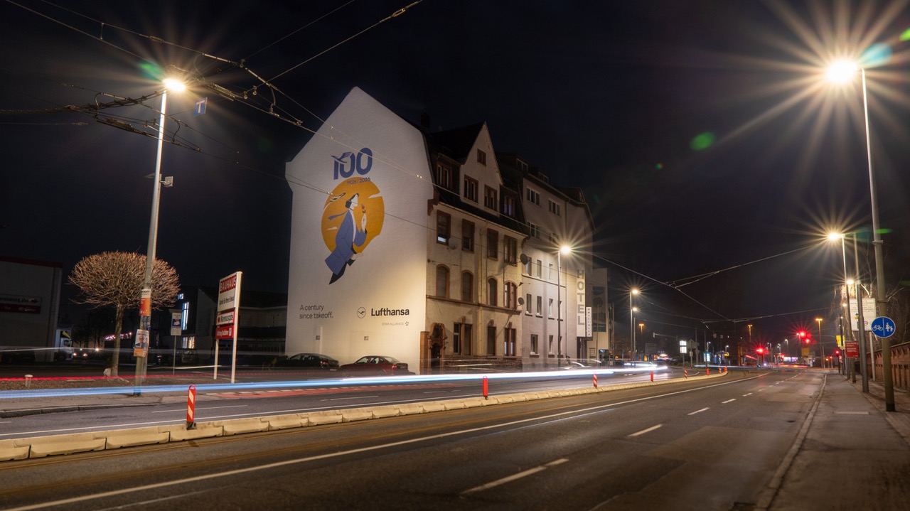 Beleuchtetes Lufthansa Jubiläums-Mural an einer Hausfassade in Frankfurt bei Nacht mit Lichtspuren vorbeifahrender Autos.
