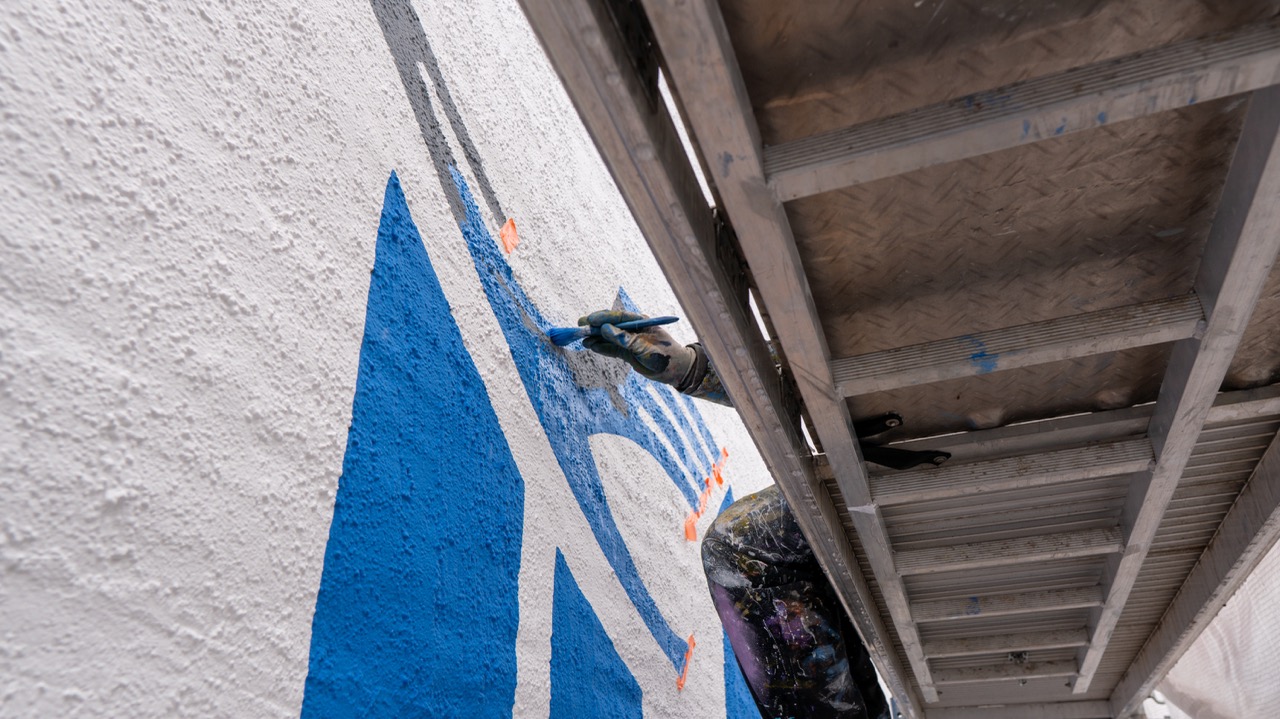 Blick von unten auf einen Künstler beim Ausmalen großer blauer Formen an einer Hausfassade.