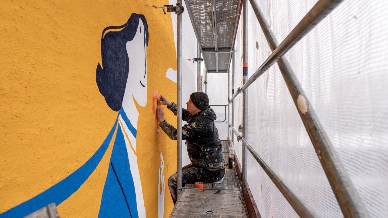 Künstler arbeitet auf einem Gerüst an einem gelb-blauen Lufthansa Jubiläums-Mural mit stilisierter Figur in Frankfurt.