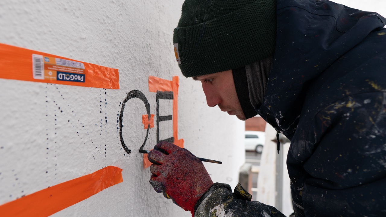 Nahaufnahme eines Artists beim präzisen Ausmalen schwarzer Schriftzüge auf einer weißen Fassadenwand.