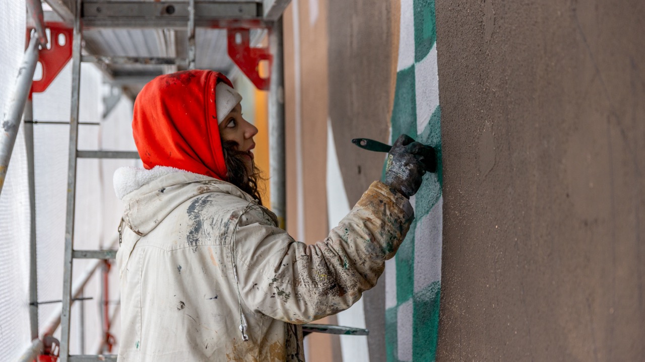 Künstlerin im roten Hoodie arbeitet auf einem Gerüst an den ersten Details eines großformatigen Lufthansa-Murals in Hamburg.
