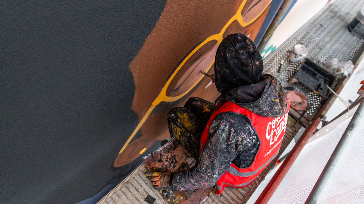 Top-Down-Aufnahme eines Künstlers auf dem Gerüst beim Ausarbeiten eines großformatigen Murals mit organischen Formen und kräftigen Farben.