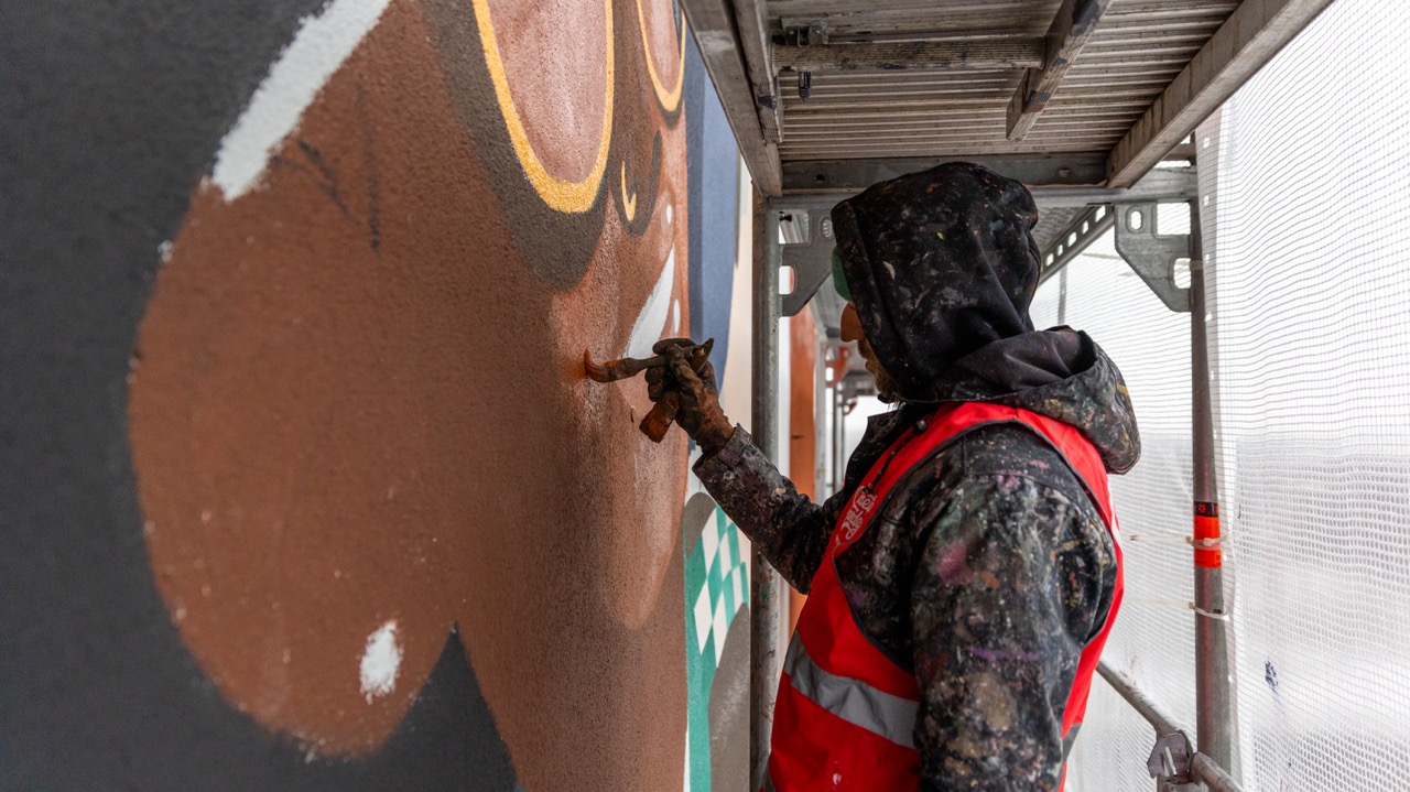Artist mit Farbrolle und Pinsel arbeitet an den Konturen eines braun-orangenen Motivs auf einer großen Hausfassade.