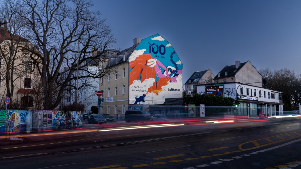 Großformatiges Lufthansa Jubiläums-Mural in München bei Abenddämmerung mit vorbeiziehenden Lichtspuren des Verkehrs.