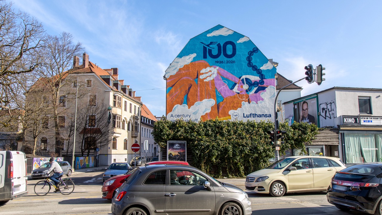 Großes Lufthansa Anniversary Mural an einer Münchner Hausfassade bei sonnigem Wetter und dichtem Stadtverkehr.