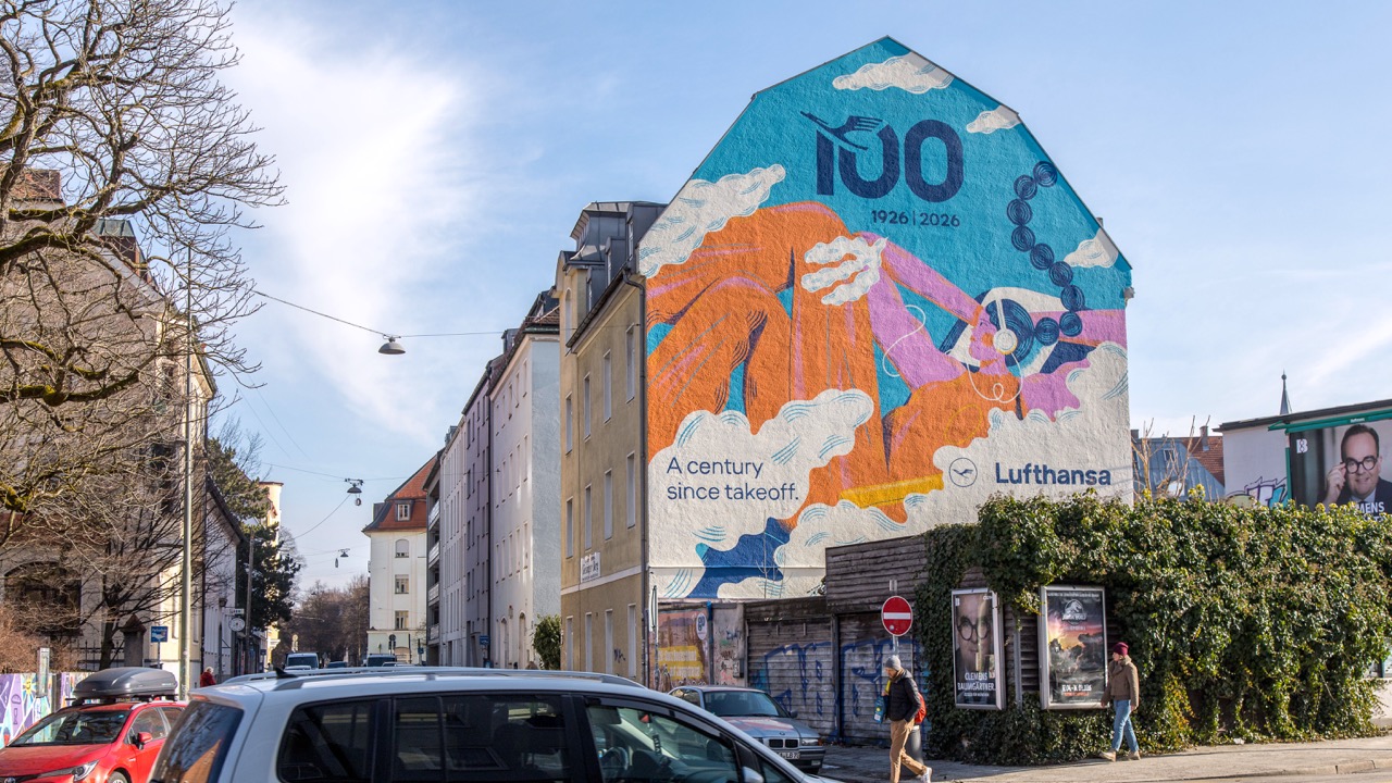 Weitwinkelaufnahme eines bunten Lufthansa-Murals an einer markanten Gebäudefassade in München.