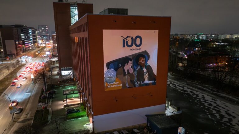 Beleuchtetes Lufthansa Anniversary Mural an einer Hamburger Hausfassade bei Nacht mit vorbeifahrendem Stadtverkehr.