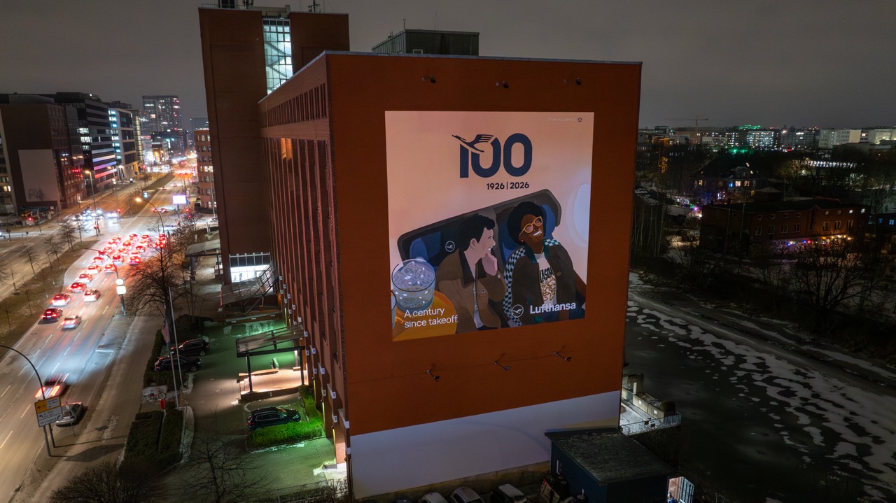 Beleuchtetes Lufthansa Anniversary Mural an einer Hamburger Hausfassade bei Nacht mit vorbeifahrendem Stadtverkehr.