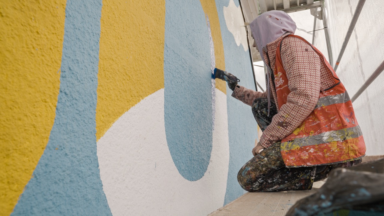 Artist malt große hellblaue und gelbe Formen auf die strukturierte Fassadenwand eines Murals.